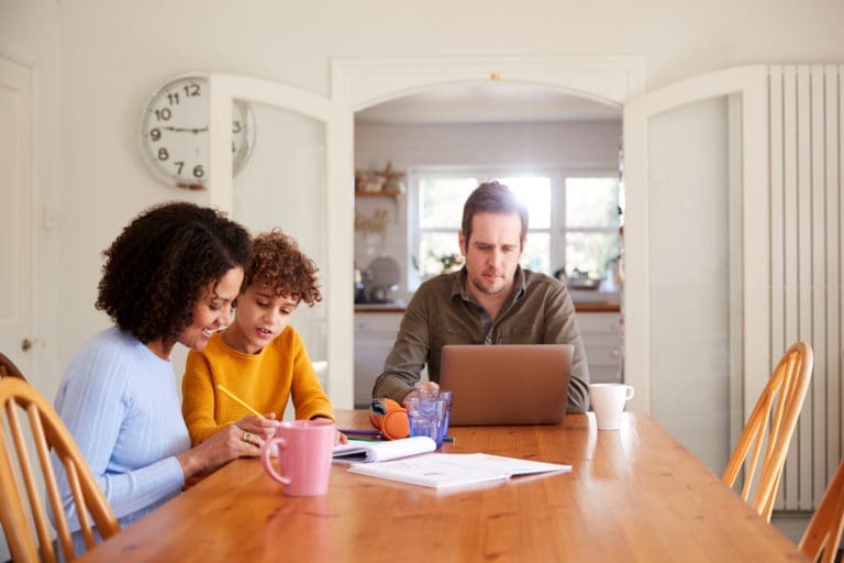 Father Works On Laptop As Mother Helps Son With Homework On Kitchen Table