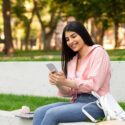 Smiling hispanic lady messaging on cellphone while relaxing in park or college campus outdoors, student girl sitting on bench and browsing internet on mobile phone, free space