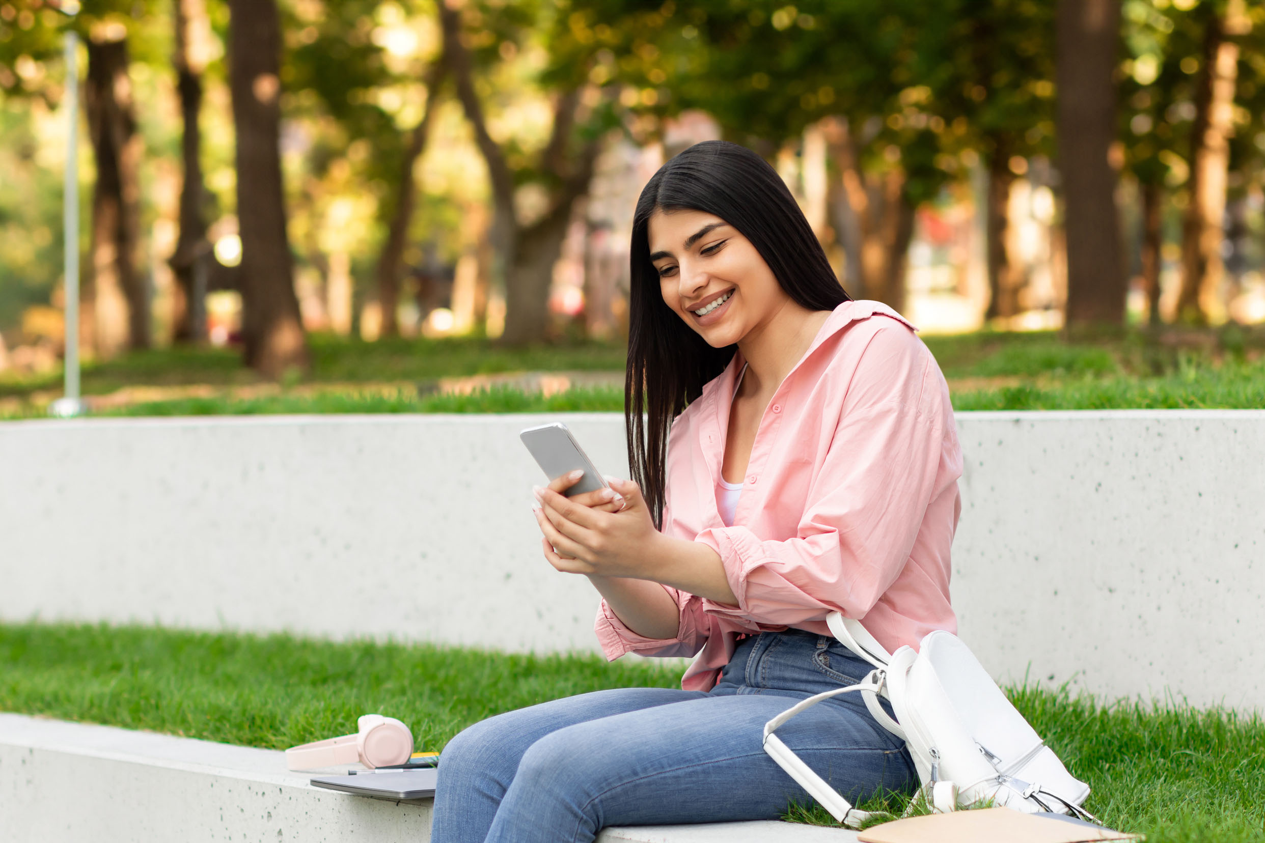 Smiling hispanic lady messaging on cellphone while relaxing in park or college campus outdoors, student girl sitting on bench and browsing internet on mobile phone, free space